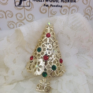 May include: Gold tone Christmas tree brooch with red and green rhinestones. The brooch is in the shape of a stylized Christmas tree with a decorative base. The text "Everbest Hollywood, Florida Pat. Pending" is visible on the box.