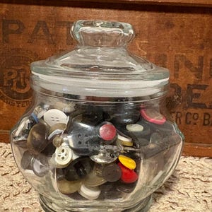 May include: A clear glass jar filled with a variety of buttons in different colors and sizes. The jar has a lid and sits on a lace doily. A wooden box is in the background.