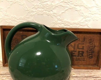 May include: A dark green ceramic pitcher with a round body and a handle. The pitcher is sitting on a wooden surface.