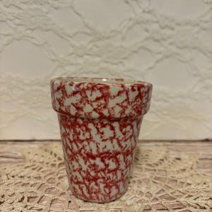 May include: A small, ceramic flower pot with a speckled red and white pattern. The pot has a slightly flared rim and a cylindrical shape. The background is a cream-colored lace doily and a white, textured surface.
