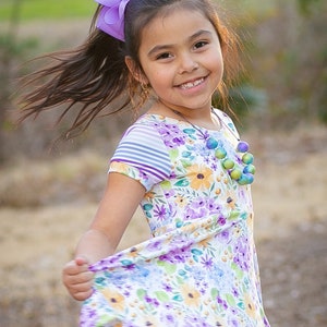 May include: A young girl with long brown hair wears a white dress with a floral pattern. She has a purple bow in her hair and a colorful beaded necklace.