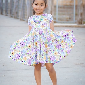 May include: A young girl wearing a floral print dress with a purple bow in her hair and yellow shoes. She is standing on a sidewalk.