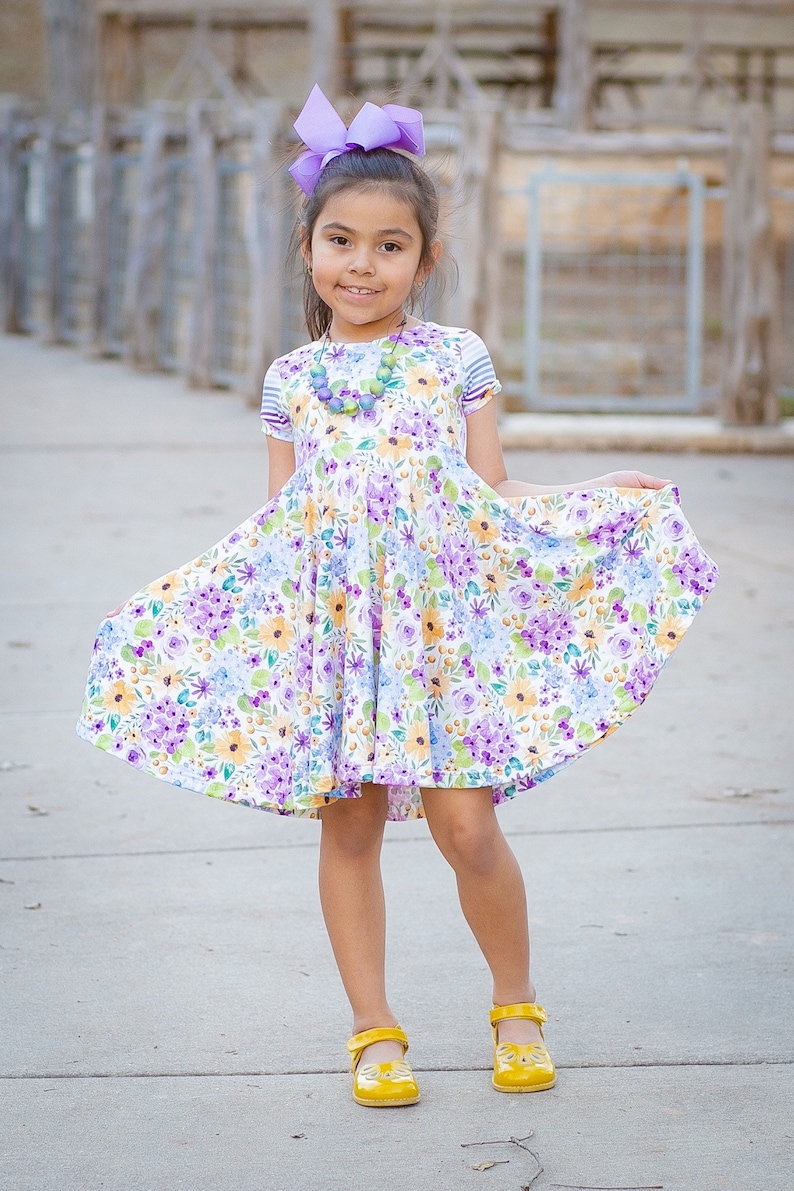May include: A young girl wearing a floral print dress with a purple bow in her hair and yellow shoes. She is standing on a sidewalk.