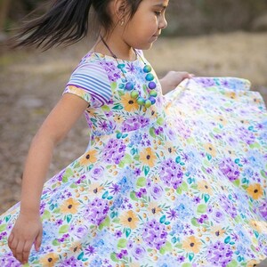 May include: A young girl wearing a white dress with a floral print in shades of purple, blue, and yellow. She is wearing a purple bow in her hair and a multi-colored beaded necklace.