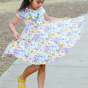 May include: A young girl wearing a white dress with a floral pattern in shades of purple, yellow, and blue. She is wearing a purple bow in her hair and a necklace with colorful beads. She is wearing yellow shoes and is twirling her dress.