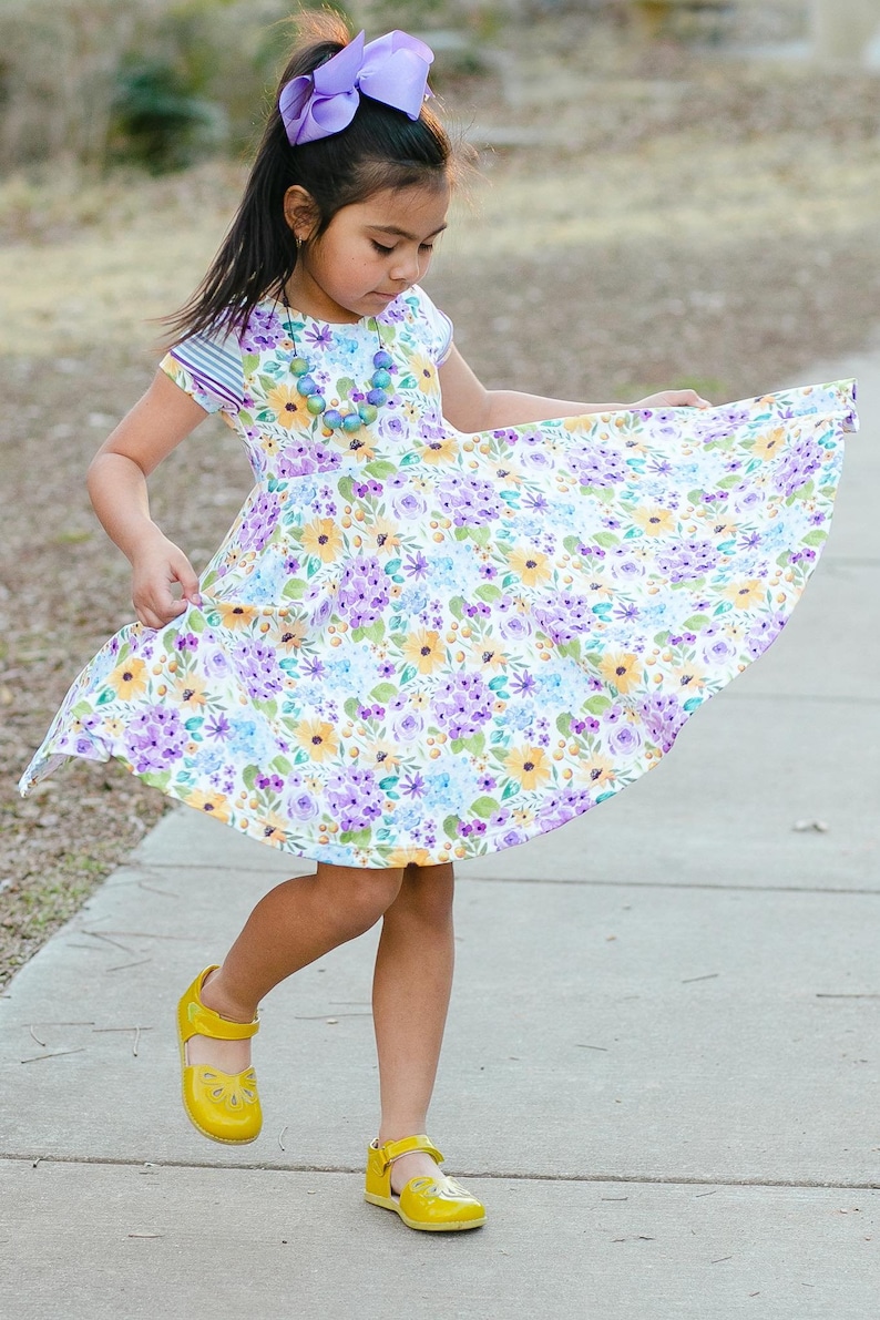 May include: A young girl wearing a white dress with a floral pattern in shades of purple, yellow, and blue. She is wearing a purple bow in her hair and a necklace with colorful beads. She is wearing yellow shoes and is twirling her dress.
