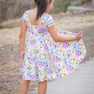 May include: A young girl wearing a white dress with a floral print in shades of purple, yellow, and blue. The dress has short sleeves and a scoop neckline. She is wearing yellow shoes and has her hair pulled back in a ponytail with a purple bow.