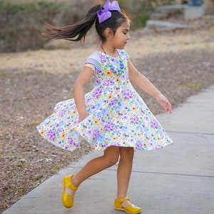 May include: A young girl wearing a white dress with a floral print, a purple bow in her hair, and yellow shoes. She is twirling on a sidewalk.