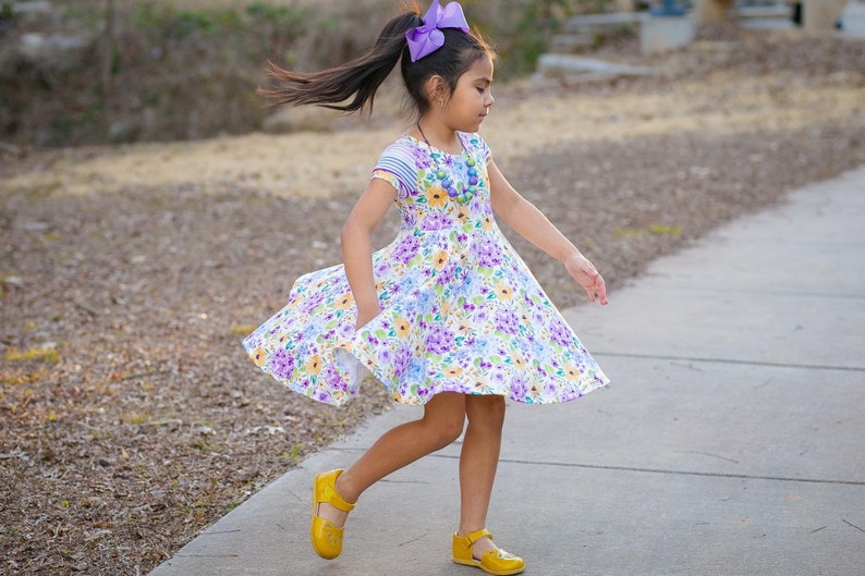 May include: A young girl wearing a white dress with a floral print, a purple bow in her hair, and yellow shoes. She is twirling on a sidewalk.