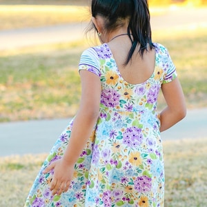 May include: A young girl wearing a white dress with a floral pattern in shades of purple, yellow, and blue. The dress has short sleeves with a striped pattern and a scoop neckline.