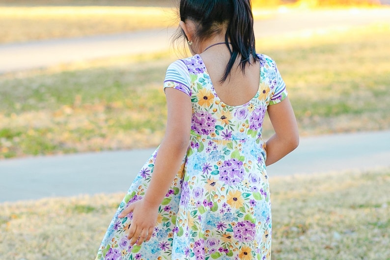 May include: A young girl wearing a white dress with a floral pattern in shades of purple, yellow, and blue. The dress has short sleeves with a striped pattern and a scoop neckline.