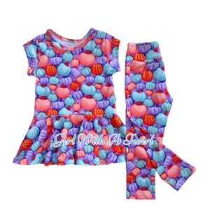 以下が含まれることがあります： 紫とピンクの半袖トップスとレギンスのセット。どちらもカラフルなカボチャのプリントが施されています。トップスには「Girl With A Twirl」の文字がプリントされています。