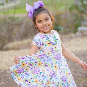 May include: A young girl wearing a floral print dress with a purple bow in her hair and a colorful beaded necklace. She is smiling and twirling in a grassy area.