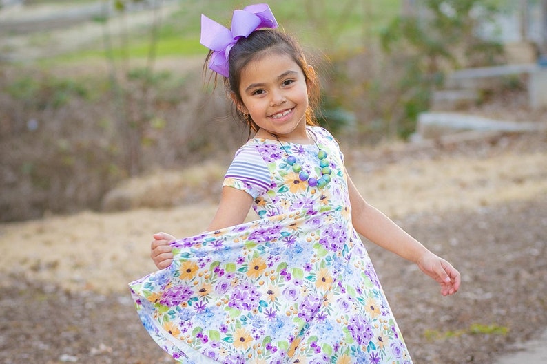 May include: A young girl wearing a floral print dress with a purple bow in her hair and a colorful beaded necklace. She is smiling and twirling in a grassy area.