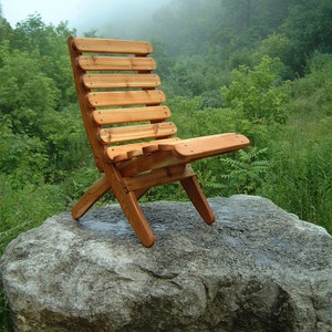 May include: A wooden folding chair with a natural wood finish sits on a large gray rock. The chair is made of slats and has a simple design. The background is a blurry image of a forest with fog.