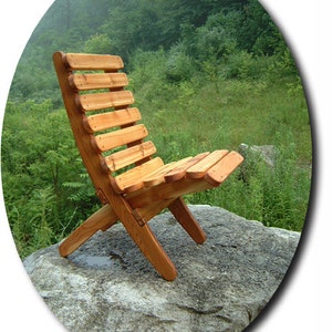 May include: A wooden folding chair with a natural finish, sitting on a large gray rock in a grassy area. The chair is made of slats of wood and has a simple, rustic design.