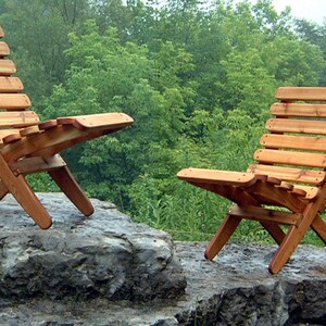 May include: Two wooden folding chairs with slatted backs and seats, made of natural cedar wood, are placed on a large gray rock. The chairs are positioned facing each other, with the backs of the chairs facing the camera. The chairs are in a natural outdoor setting with green trees in the background.