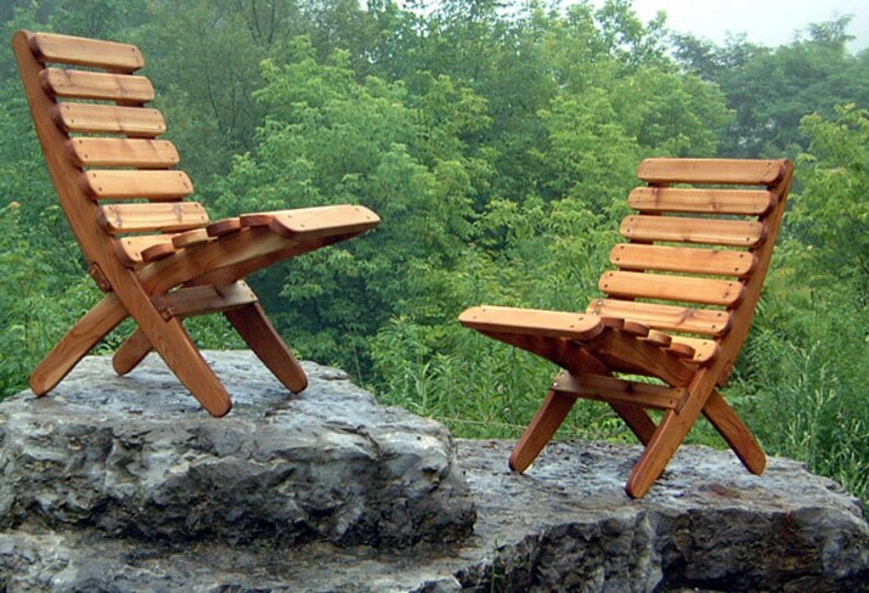 May include: Two wooden folding chairs with slatted backs and seats, made of natural cedar wood, are placed on a large gray rock. The chairs are positioned facing each other, with the backs of the chairs facing the camera. The chairs are in a natural outdoor setting with green trees in the background.
