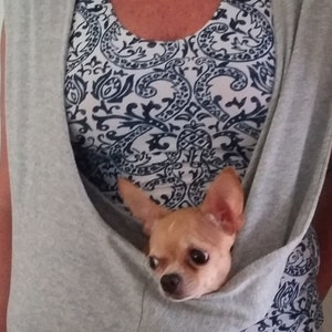 以下が含まれることがあります： 小さな犬を入れるためのポケットが内蔵されたグレーのノースリーブシャツ。小さなチワワがポケットに座ってカメラを見ている。