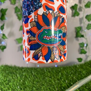 May include: A white tumbler with a colorful design featuring a blue and green alligator head with orange and white accents. The design also includes a floral pattern with orange, blue, and brown colors. The tumbler has a white background with a leopard print pattern.