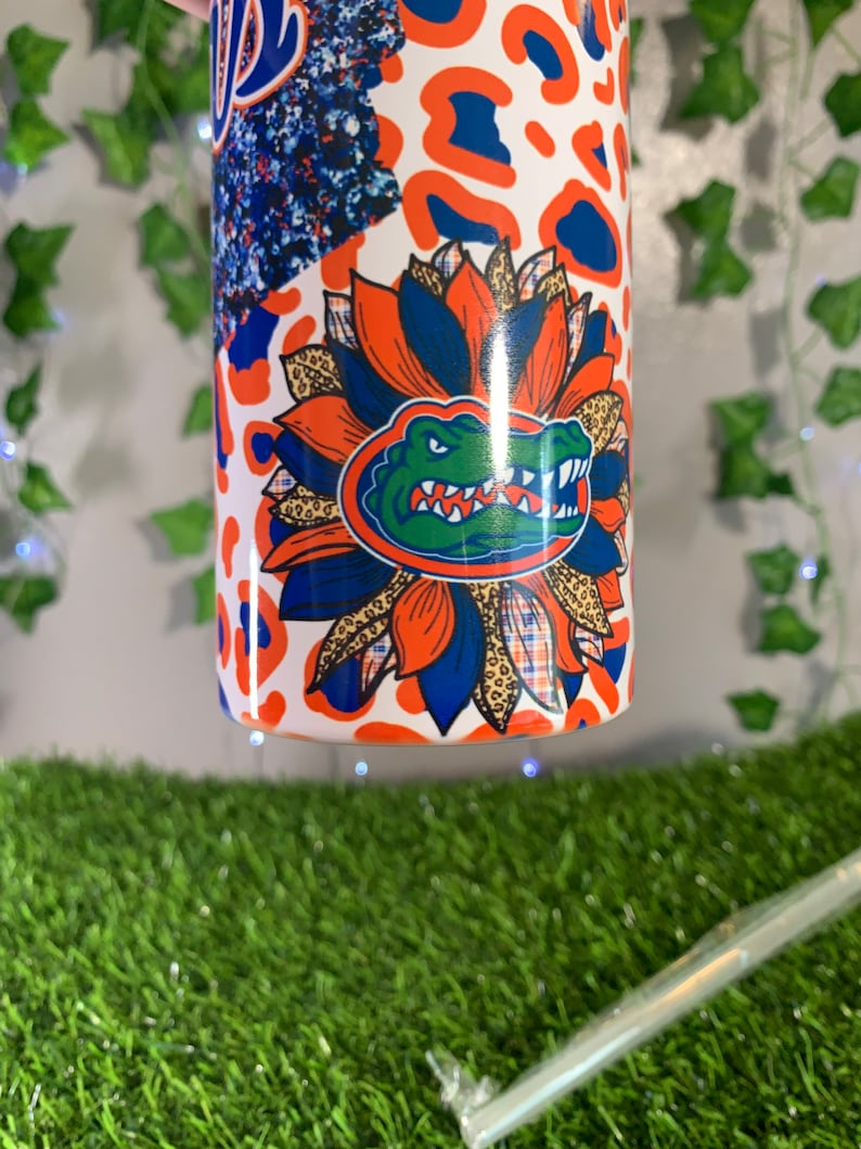 May include: A white tumbler with a colorful design featuring a blue and green alligator head with orange and white accents. The design also includes a floral pattern with orange, blue, and brown colors. The tumbler has a white background with a leopard print pattern.
