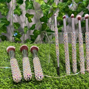 May include: A set of makeup brushes with iridescent rhinestone-covered handles and soft, pink-toned bristles. The brushes are arranged on a clear acrylic stand, with a backdrop of green foliage.