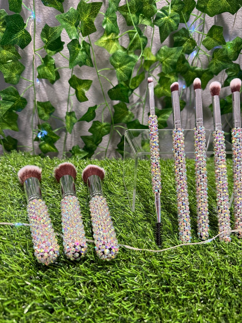 May include: A set of makeup brushes with iridescent rhinestone-covered handles and soft, pink-toned bristles. The brushes are arranged on a clear acrylic stand, with a backdrop of green foliage.