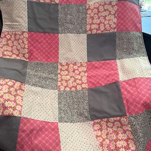 May include: A patchwork quilt featuring a variety of fabrics in a grid pattern. The quilt incorporates squares of pink floral, gray leaf, white polka dot, and solid gray fabrics. The quilt is a mix of patterns and colors.