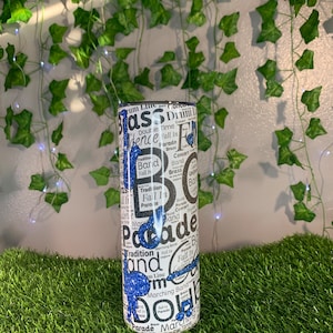 May include: A white tumbler with a blue glitter design featuring the words "Bass Drum", "Fall In", "Parade", "Marching Band", and "Double Time".
