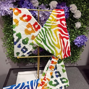 May include: A display of colorful headbands with rainbow zebra and leopard prints. The headbands are displayed on a gold metal stand against a backdrop of purple and green foliage. The headbands are made of fabric.