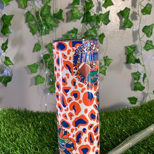 May include: A white tumbler with a blue and orange leopard print design. The tumbler features the text "Gator Nation" and "Florida" with a cartoon alligator and a football.