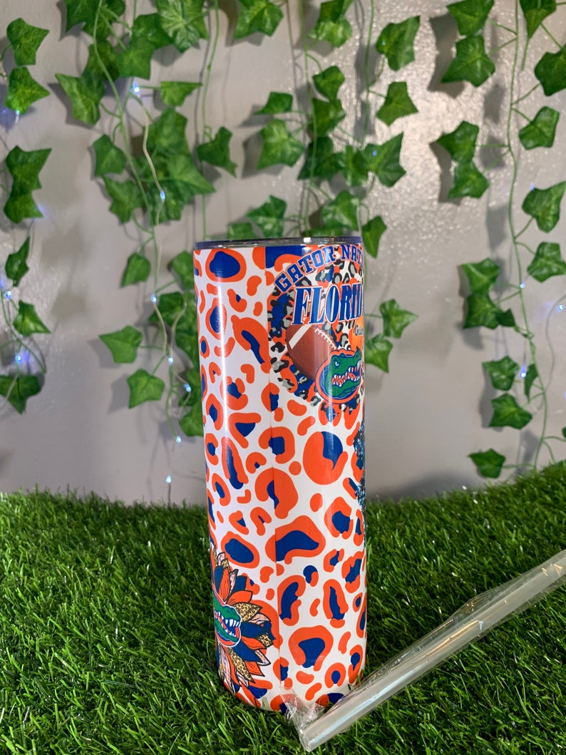 May include: A white tumbler with a blue and orange leopard print design. The tumbler features the text "Gator Nation" and "Florida" with a cartoon alligator and a football.
