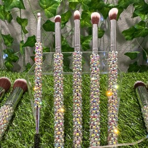 May include: A set of makeup brushes with clear, crystal-like embellishments on the handles. The bristles are various shades of brown and white. The brushes are displayed on a clear stand against a backdrop of green foliage.