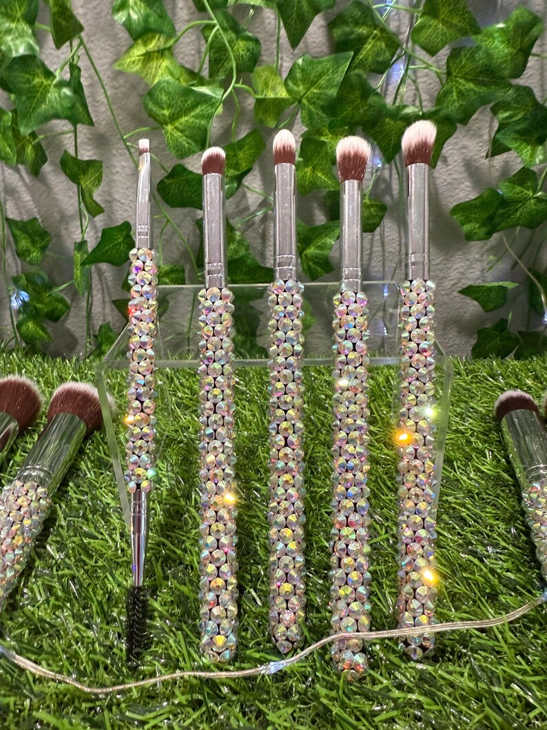 May include: A set of makeup brushes with clear, crystal-like embellishments on the handles. The bristles are various shades of brown and white. The brushes are displayed on a clear stand against a backdrop of green foliage.