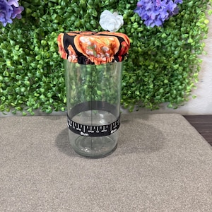 May include: A clear glass jar with a black and white band that reads "Sun" and "Mon". The jar is topped with a fabric lid featuring a colorful pattern of oranges and other food items. The background is a green wall with purple and white flowers.