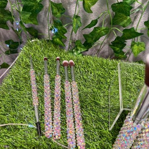 May include: A collection of makeup brushes with iridescent, crystal-like handles. The brushes have silver metal stems and soft, pink bristles. One brush is a mascara wand. The brushes are arranged on a bed of green artificial grass.