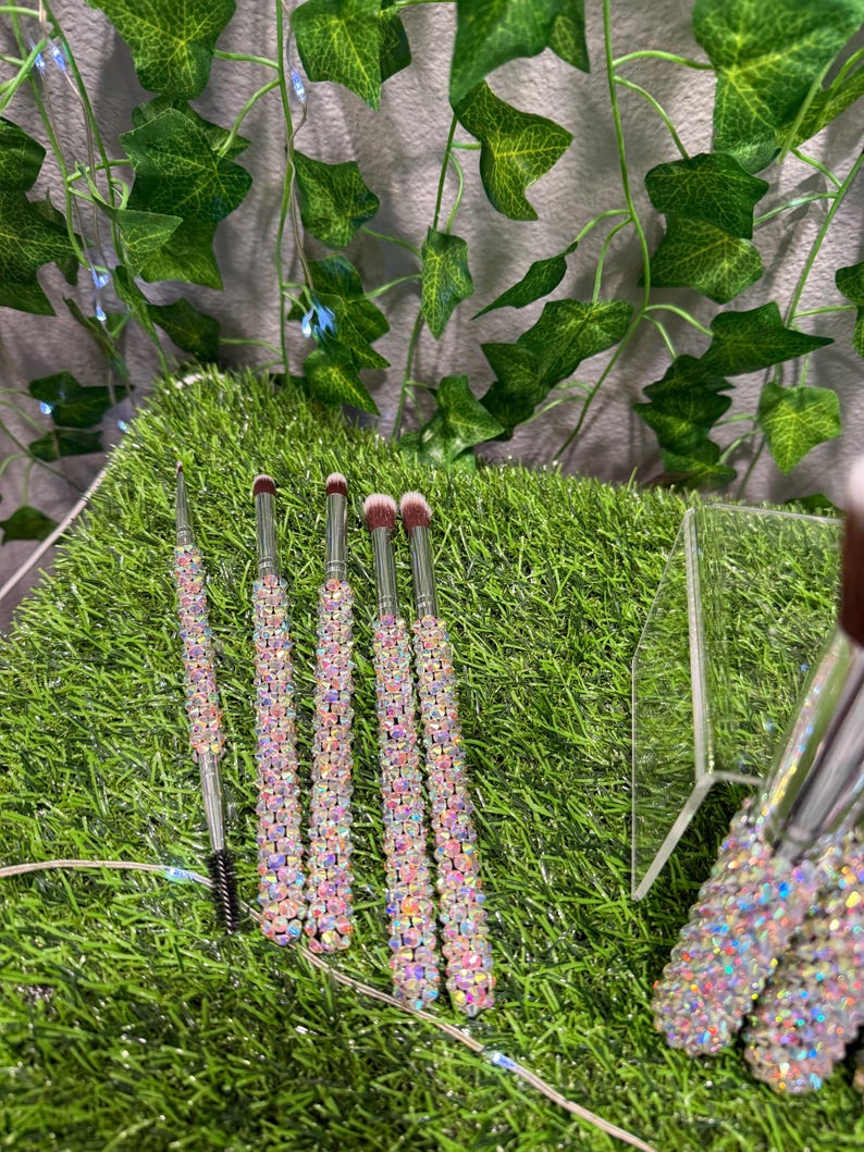 May include: A collection of makeup brushes with iridescent, crystal-like handles. The brushes have silver metal stems and soft, pink bristles. One brush is a mascara wand. The brushes are arranged on a bed of green artificial grass.