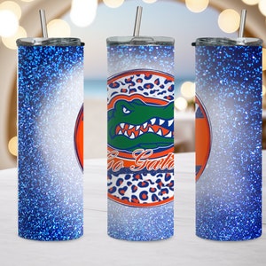 May include: Three stainless steel tumblers with blue glitter designs. The center tumbler features a green alligator with the words "Go Gators" in a white circle with orange and black leopard print accents.