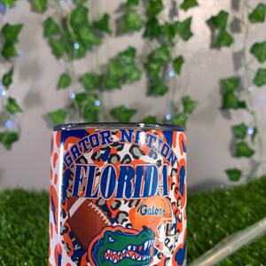 May include: A white tumbler with an orange, blue, and gray Gator Nation Florida design. The design features a gator, a football, and a helmet with the word "Gators" on it.