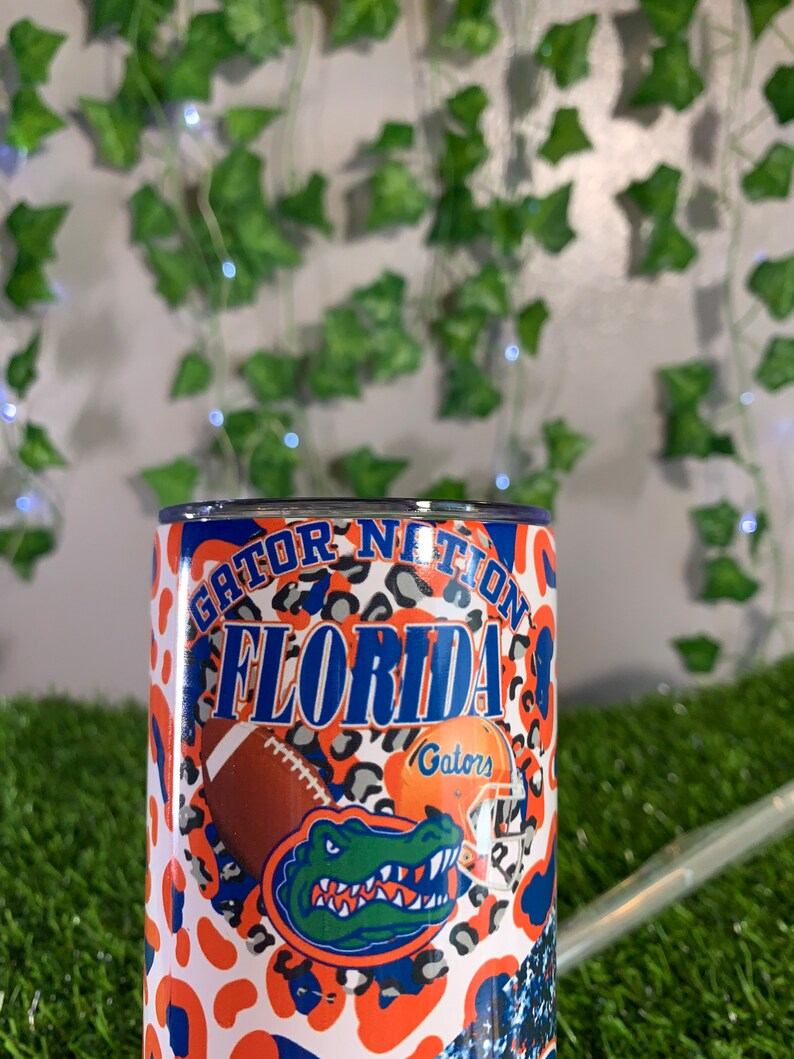 May include: A white tumbler with an orange, blue, and gray Gator Nation Florida design. The design features a gator, a football, and a helmet with the word "Gators" on it.
