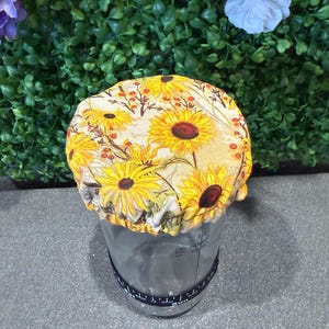 May include: A clear glass jar with a black band and a yellow sunflower fabric lid. The lid features a repeating pattern of sunflowers and berries. The jar is on a gray surface.