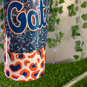 May include: A white tumbler with a blue and orange leopard print design. The top half of the tumbler features a blue glitter background with the word "Gators" in a blue script font.