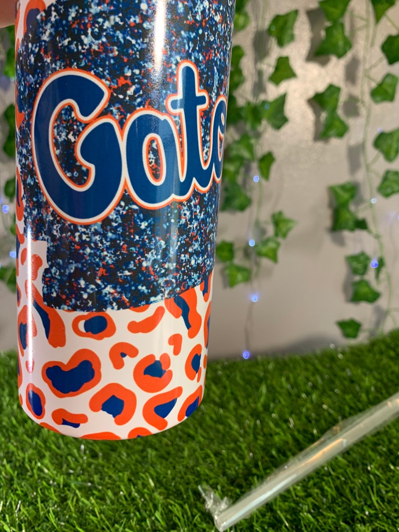 May include: A white tumbler with a blue and orange leopard print design. The top half of the tumbler features a blue glitter background with the word "Gators" in a blue script font.