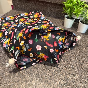 May include: Black drawstring bag with a colorful ramen pattern. The design includes illustrations of ramen bowls, eggs, shrimp, and flowers. The bag has a white drawstring closure with wooden bead accents. The bag is open, revealing the interior.