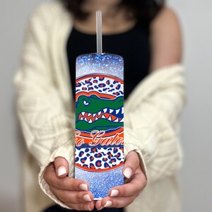 May include: A blue and white tumbler with a glitter finish and a University of Florida Gators logo. The logo features an alligator with the words "Go Gators" in orange and blue.