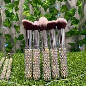 May include: A set of makeup brushes with silver handles encrusted with iridescent rhinestones. The brush heads are a mix of light and dark brown, and white. The brushes are arranged on a bed of green grass.