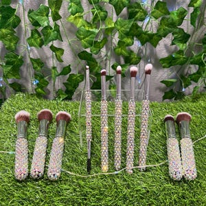 May include: A set of makeup brushes with sparkling, iridescent handles. The brushes have pink and brown bristles. The handles are encrusted with small, shiny crystals. The brushes are arranged on a bed of green artificial grass.