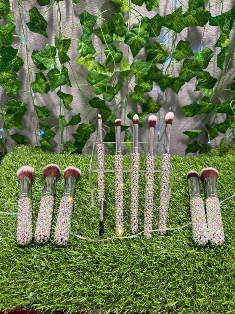 May include: A set of makeup brushes with sparkling, iridescent handles. The brushes have pink and brown bristles. The handles are encrusted with small, shiny crystals. The brushes are arranged on a bed of green artificial grass.