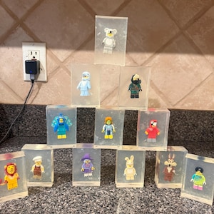 May include: A collection of rectangular clear soap bars, each containing a unique plastic minifigure. The figures include a lion, chef, witch, rabbit, wolf, peacock, space character, parrot, shark, koala, and others, arranged in a pyramid formation.