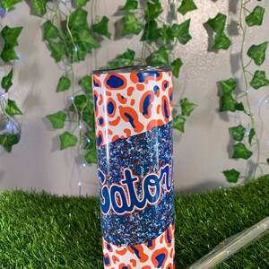 May include: A white, blue, and orange stainless steel tumbler with a leopard print design and the word "Gator" in blue lettering.